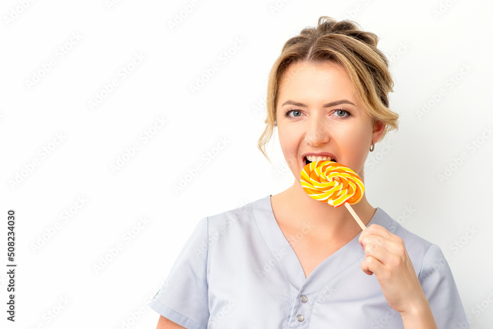 Portrait of a beautiful young caucasian beautician wearing a medical shirt bites a lollipop on a white background
