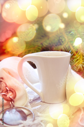 Blurry holiday scene of white cup of hot cocoa and white fluffy plaid against blurred background with beautiful Christmas lights of bokeh.