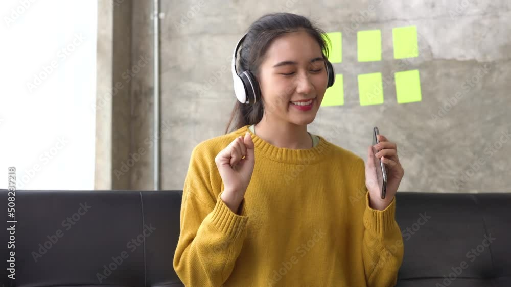 Happy asian woman relaxing and using overhead headphones to listen to music from smart phone sitting on sofa. Happy asian woman relaxing and using overhead headphones to listen to music from smart phone sitting on sofa.