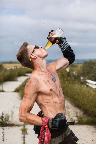 male vandal with dirty naked torso drinks beer, against backdrop of an overgrown abandoned road