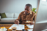 African senior man sitting at home and looking at bills he has to pay. He is paying it online over a PC computer. Serious senior man sitting in living room manage budget received invoice