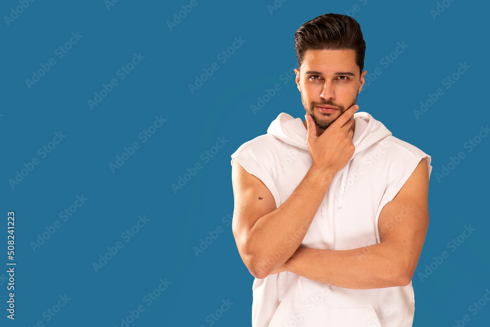 Naklejka premium Fashionable young handsome man posing in white tank top with hood on blue studio background. Bearded guy looking at the camera.