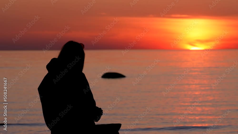 Young woman with sun glasses silhouette by water during sunset. Nodding in talking to someone. Eating. Bird flying. Young woman with sun glasses silhouette by water during sunset. Nodding in talking to someone. Eating. Bird flying.