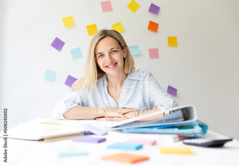Portrait of pretty young blonde woman with beautiful laugh sitting at office table in front of white wall with colorful sticky notes