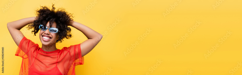 joyful african american woman in red top and blue sunglasses posing isolated on yellow, banner.