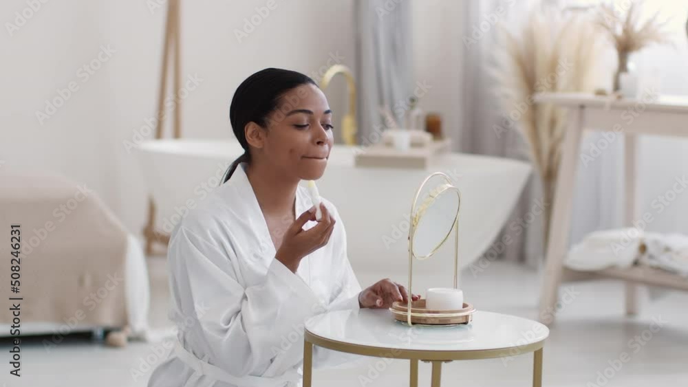 Young pretty african american lady wearing bathrobe applying hygienic lipstick on lips, looking at mirror, empty space Young pretty african american lady wearing bathrobe applying hygienic lipstick on lips, looking at mirror, empty space