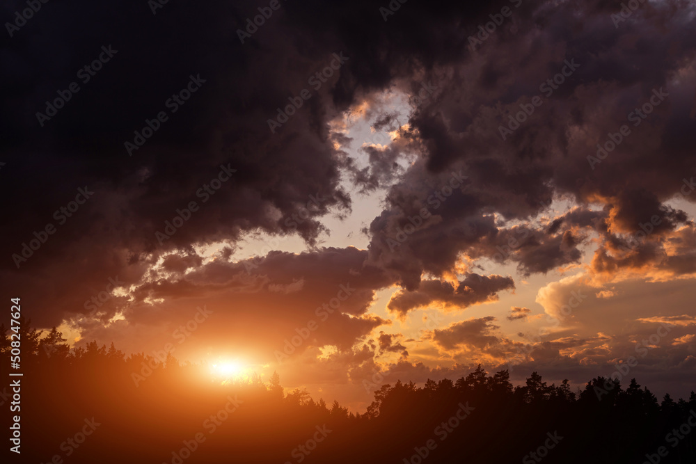 Epic dramatic sunset on storm sky with dark grey clouds, orange yellow sun and sunlight above black silhouette trees in forest