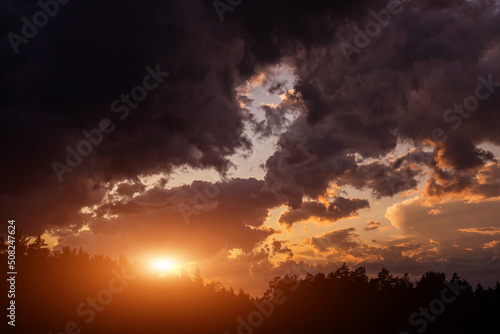 Epic dramatic sunset on storm sky with dark grey clouds, orange yellow sun and sunlight above black silhouette trees in forest