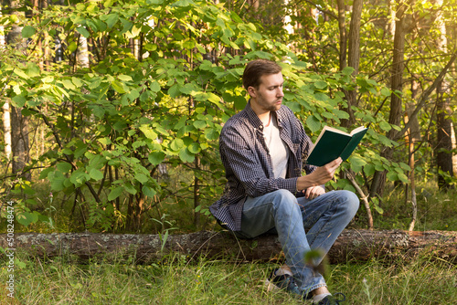 Young man alone resting and reading book in summer green forest outdoors in nature in sunlight