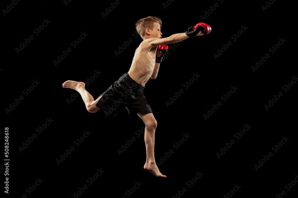 Studio shot of preschool boy, cute kid in sports shots and gloves training alone on black background. Sport, education, action, motion concept.