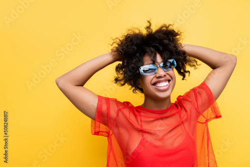 excited african american woman in red stylish top and sunglasses touching wavy hair isolated on yellow.