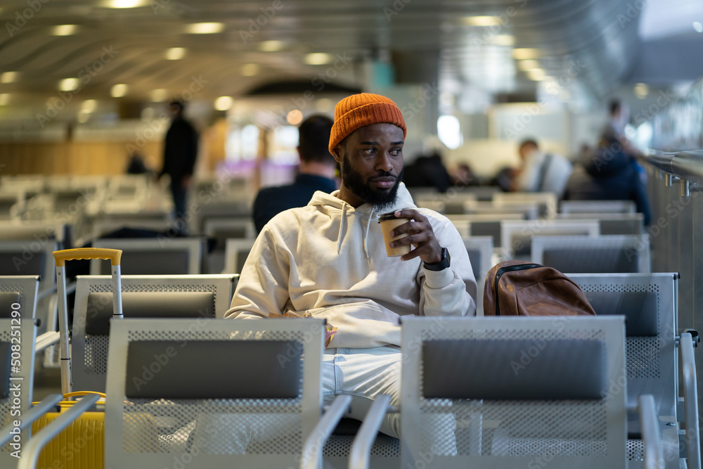 Young African American traveler man drinking coffee and eating sandwich while waiting for flying at airport terminal. Trendy black hipster guy satisfies hunger with a coffee and sandwich. Young African American traveler man drinking coffee and eating sandwich while waiting for flying at airport terminal. Trendy black hipster guy satisfies hunger with a coffee and sandwich.