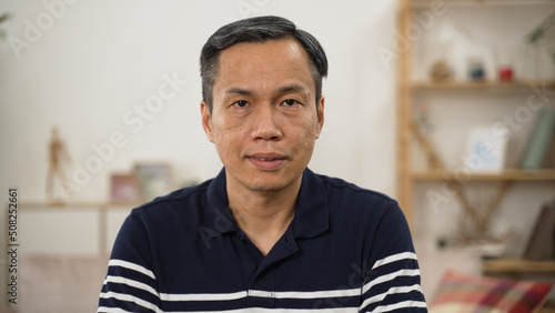 half length portrait of a grey haired Asian male pensioner in casual wear looking at camera with serious face expression on home background
