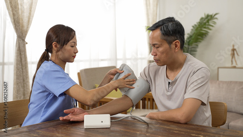 Chinese woman care attendant checking senior man’s blood pressure using a digital sphygmomanometer during home visit. domiciliary care for elderly people concept