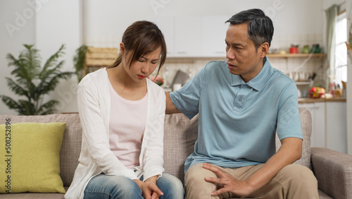 caring asian retired father consoling frustrated daughter and patting on her back in living room at home. the depressed woman cries and buries her face in hands while listening