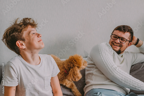 close-up portrait dad and son teenager generation Z laugh very cheerfully and provocatively at home on the couch, between them sits a pet red poodle dog