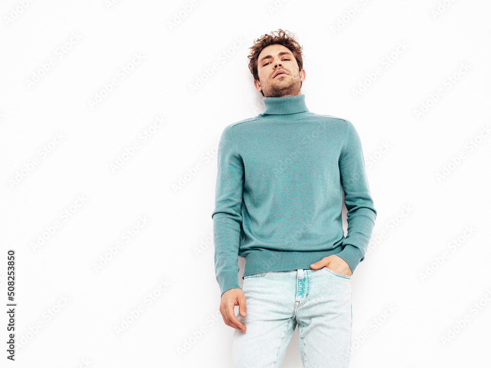 Portrait of handsome confident model. Sexy stylish man dressed in blue sweater and jeans. Fashion hipster male with curly hairstyle posing near white wall in studio. Isolated Portrait of handsome confident model. Sexy stylish man dressed in blue sweater and jeans. Fashion hipster male with curly hairstyle posing near white wall in studio. Isolated