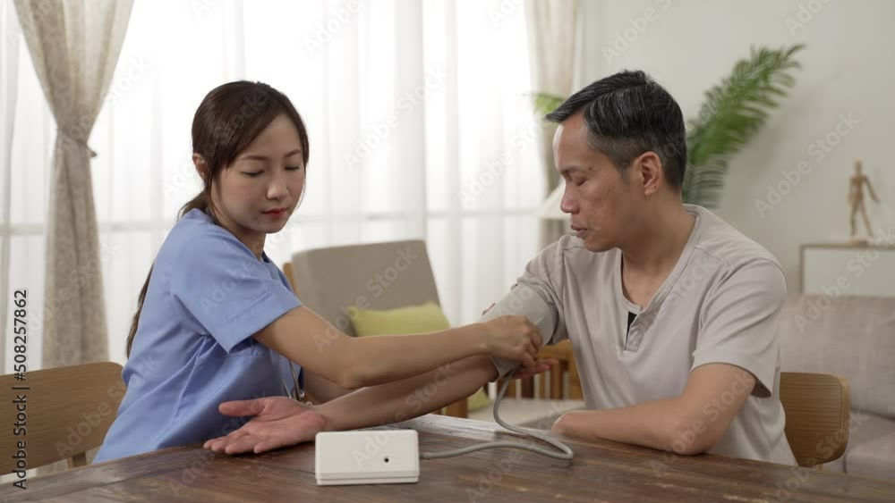 Asian female personal aide measuring blood pressure for the male elderly senior at home. she puts cuff on his upper arm and operates the device, waiting for result Asian female personal aide measuring blood pressure for the male elderly senior at home. she puts cuff on his upper arm and operates the device, waiting for result