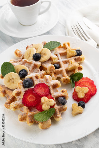 Belgian waffles with strawberries and bananas on a white plate, in the background, a cup of tea and cutlery.
