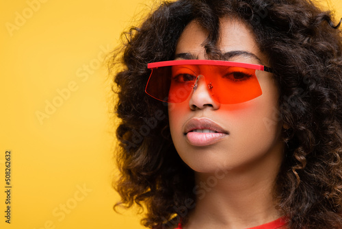 portrait of brunette woman in fashionable sunglasses looking at camera isolated on yellow.