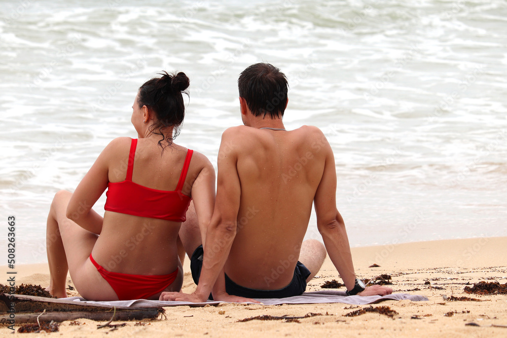 Couple sitting on a sand on sea background. Man and woman in red bikini together, romantic holiday and beach vacation Couple sitting on a sand on sea background. Man and woman in red bikini together, romantic holiday and beach vacation
