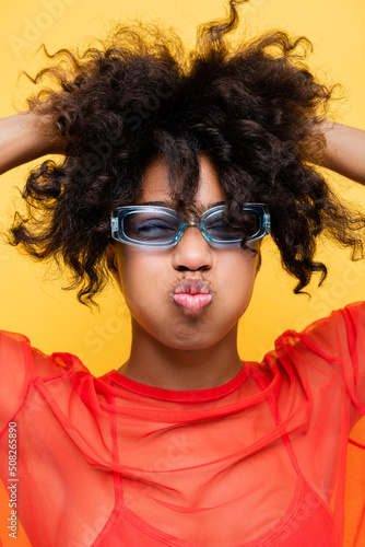 african american woman in trendy sunglasses pouting lips and touching wavy hair isolated on yellow.