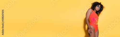 curly african american woman in trendy sunglasses and swimsuit on yellow background, banner. © LIGHTFIELD STUDIOS