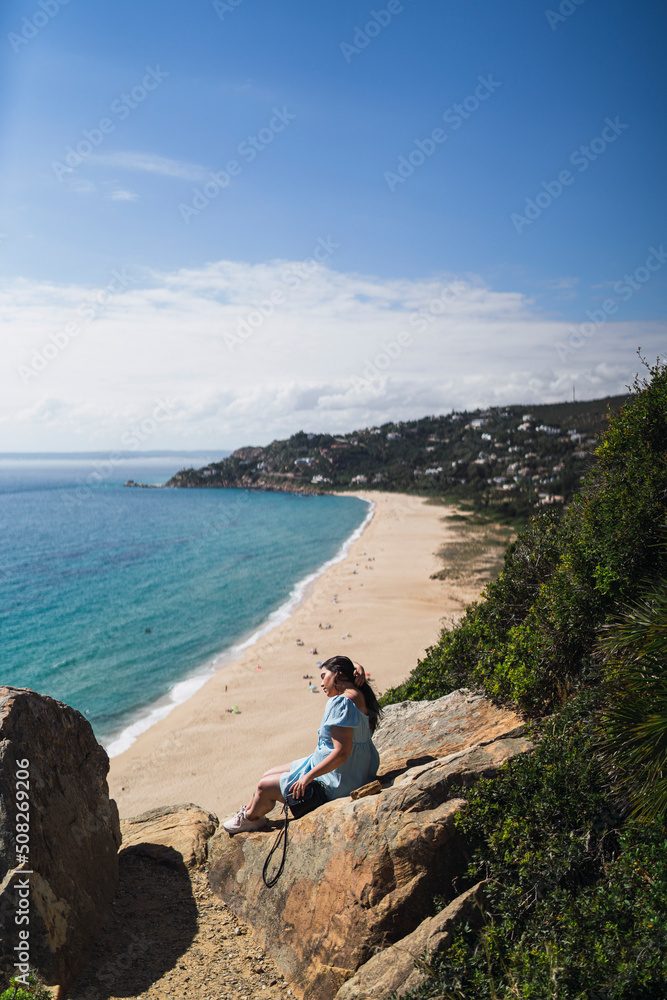 Chica joven en roca observando paisaje de playa con gente realizando parapente delante suya