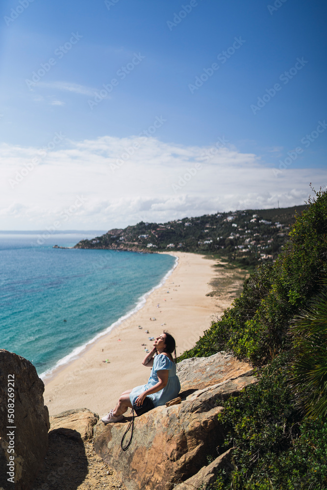Chica joven en roca observando paisaje de playa con gente realizando parapente delante suya