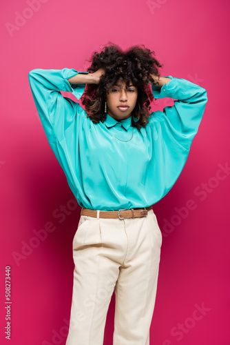 trendy african american woman in turquoise blouse touching curly hair isolated on pink.