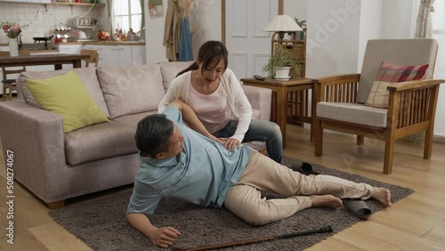 Asian senior father suffering pain from a fall and lying on living room floor with a stick beside at home. Her adult daughter quickly comes over to help and calls 911 on cellphone