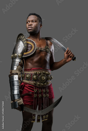 Portrait of isolated on gray gladiator of african descent dressed in light armor holding two swords.