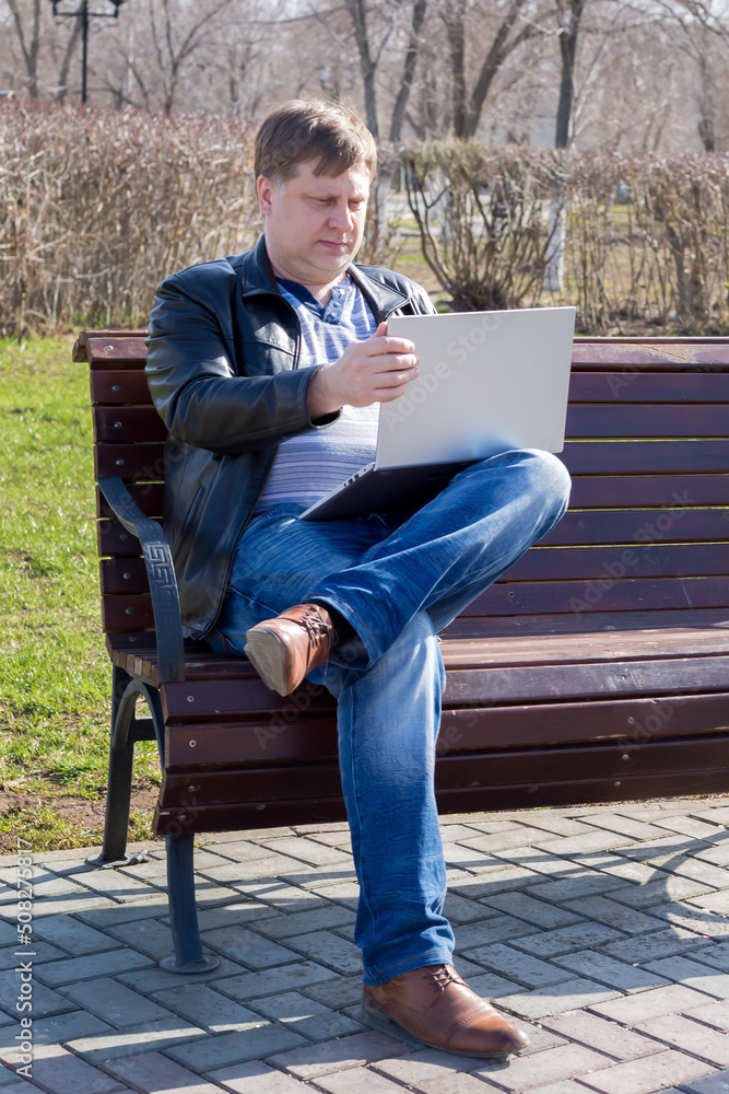 Adult male freelancer in a leather jacket works at a computer in the park sitting bench on a sunny spring day.
