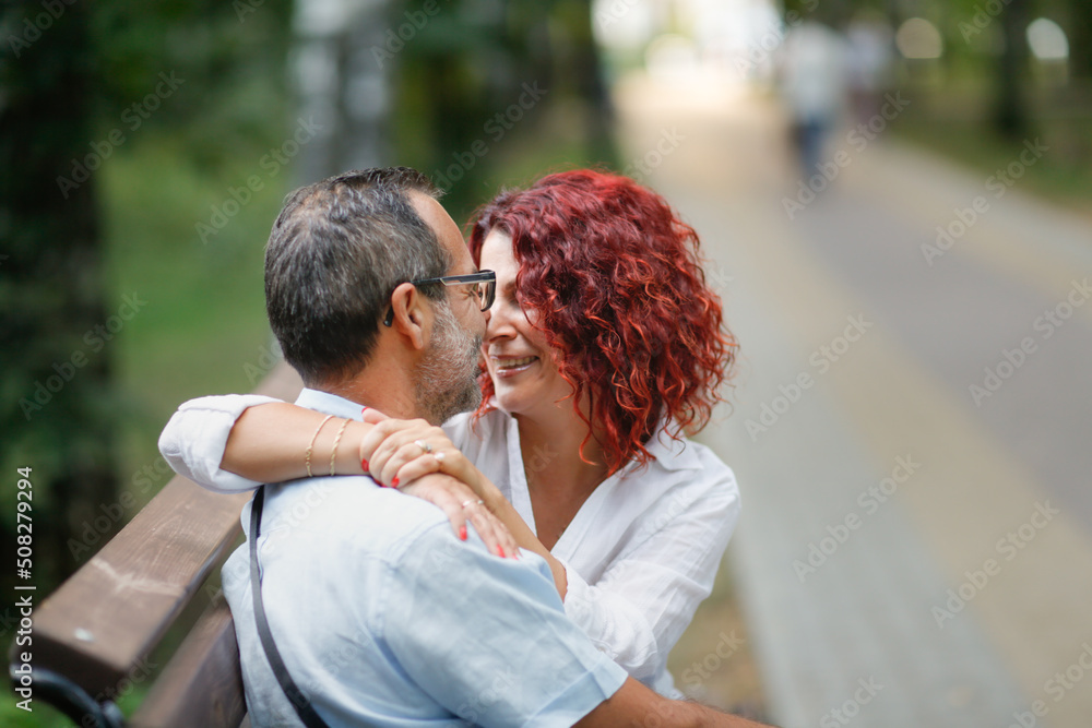red-haired pretty woman hugs slender man with glasses. Cute middle aged european couple on park bench. red-haired pretty woman hugs slender man with glasses. Cute middle aged european couple on park bench.