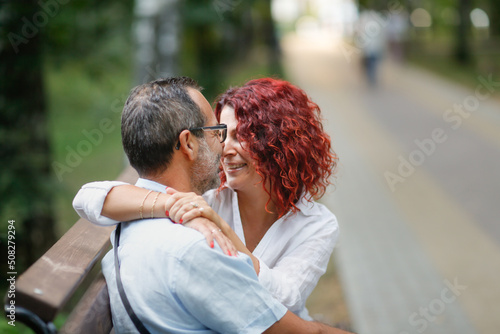 red-haired pretty woman hugs slender man with glasses. Cute middle aged european couple on park bench.