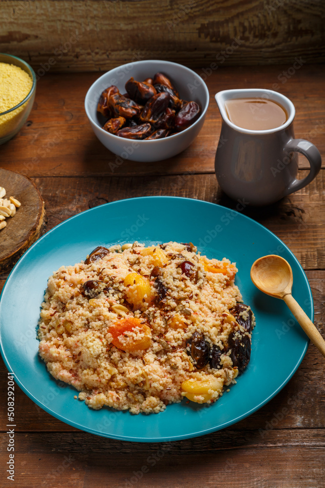 A large dish of couscous on a wooden table next to dates nuts and a jug of fruit broth.