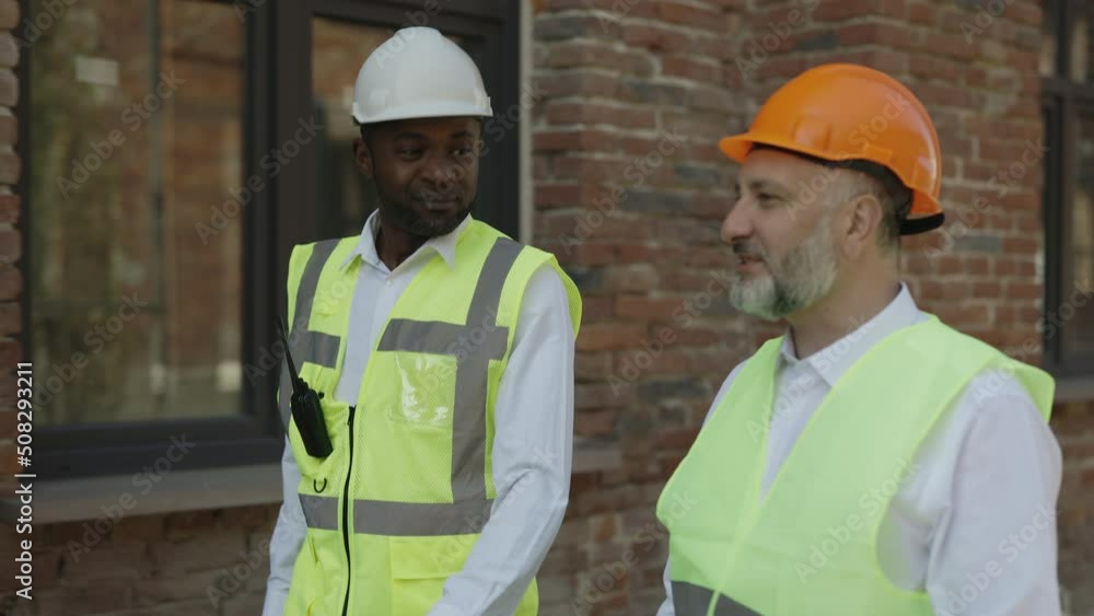 Construction engineer and foreman discussing work plan while walking on building site. Two smiling multicultural colleagues wearing reflective vests and protective helmets. Construction engineer and foreman discussing work plan while walking on building site. Two smiling multicultural colleagues wearing reflective vests and protective helmets.