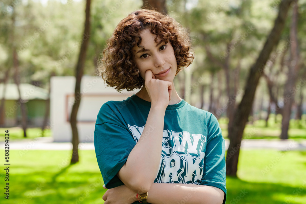 Fototapeta premium Young sad redhead woman wearing green t-shirt standing on city park, outdoors touching chin and looking at the camera, thoughtful, lost in thoughts consider idea. Making important choice.