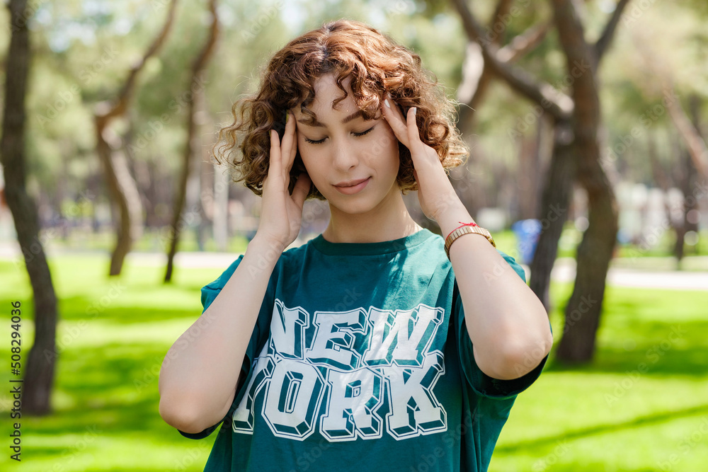 Young stressed redhead woman wearing green tee standing on city park, outdoors holding her head with hands trying to remember something or having headache. Suffering from pain.