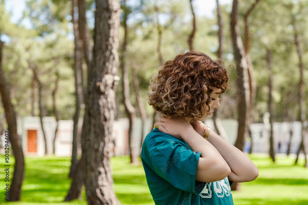 Side view of young redhead woman standing on city park, outdoors feeling hurt joint shoulder back pain ache, fibromyalgia concept, close up rear view. Health care and medical concept. Copy space. Side view of young redhead woman standing on city park, outdoors feeling hurt joint shoulder back pain ache, fibromyalgia concept, close up rear view. Health care and medical concept. Copy space.