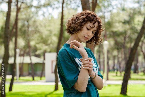 Side view of sad young woman wearing green t-shirt standing on city park  outdoors holding her painful wrist. Suffering pain on hands and fingers  arthritis inflammation.
