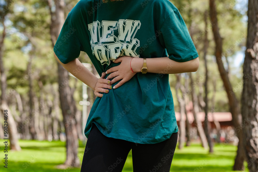 Cropped view of young sportive woman wearing green t-shirt and black legging standing on city park, outdoors with hand on stomach because indigestion, painful illness feeling unwell. Ache concept. Cropped view of young sportive woman wearing green t-shirt and black legging standing on city park, outdoors with hand on stomach because indigestion, painful illness feeling unwell. Ache concept.