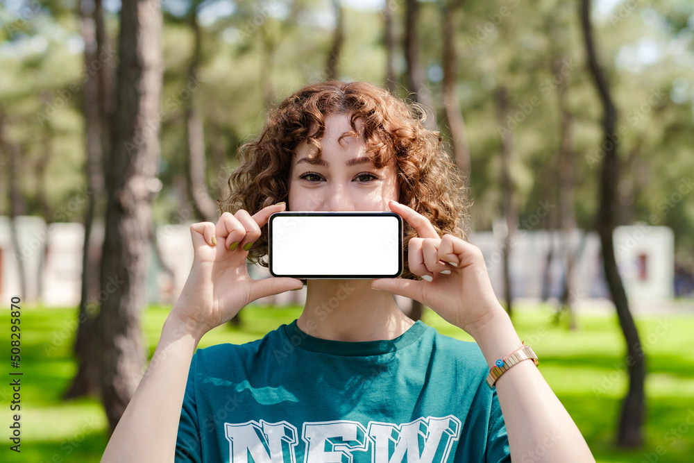 Young redhead woman wearing green t-shirt standing on city park, outdoors covering mouth with blank mockup white screen smartphone. Digital and social media concepts.