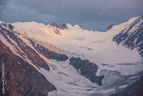 Dramatic aerial view to high snow mountain range in early morning at dawn. Awesome scenery with sunlit snow mountains in cloudy sky at sunrise. Scenic landscape with large glacier in sunrise colors. © Daniil