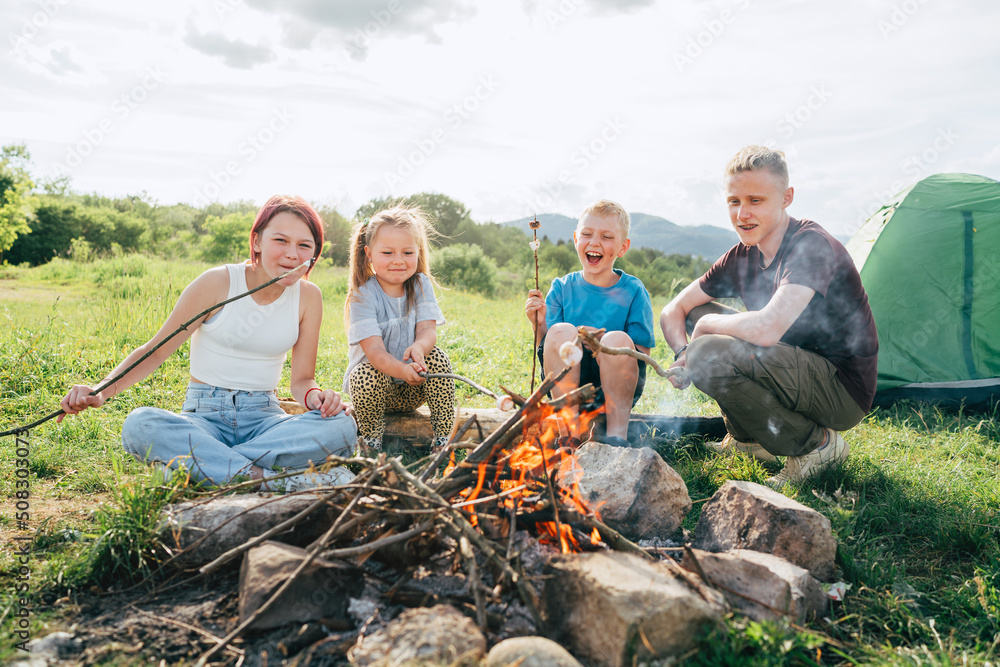 Obraz premium Boys and girls kids cheerfully laughing and roasting marshmallows and sausages on sticks over campfire flame near the green tent. Outdoor active time spending or camping in Nature concept.