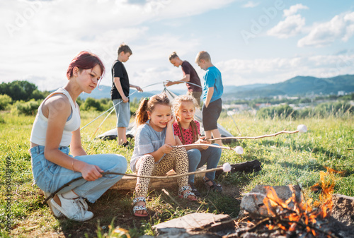 Three sisters sitting near campfire, smiling and roasting marshmallows and candies on sticks over while brothers set up the green tent. Happy family outdoor picnic camping activities concept.