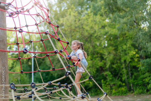 A happy little girl is climbing on an outdoor playground on a summer day. Blonde girl climbs to the top of the game complex