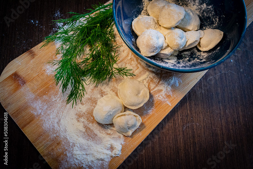 Raw dumplings in a blue plate on a cutting board and ingredients for homemade dumplings, bunches of green dill, round pepper, bay leaf. The process of making dumplings.