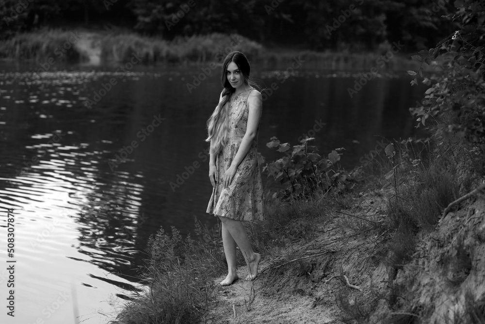 Young long-haired woman in dress standing near water in the forest in black and white Young long-haired woman in dress standing near water in the forest in black and white