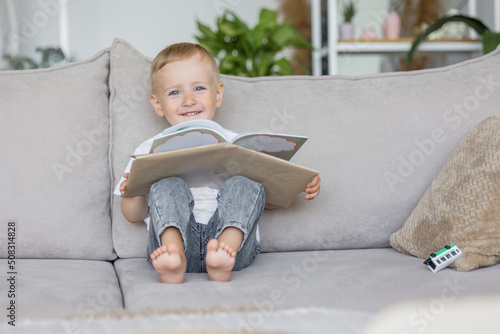 little boy is playing and reading a book at home on the sofa in the living room. a European child alone learns the alphabet and reads fairy tales. earlier development of the child at school or at home